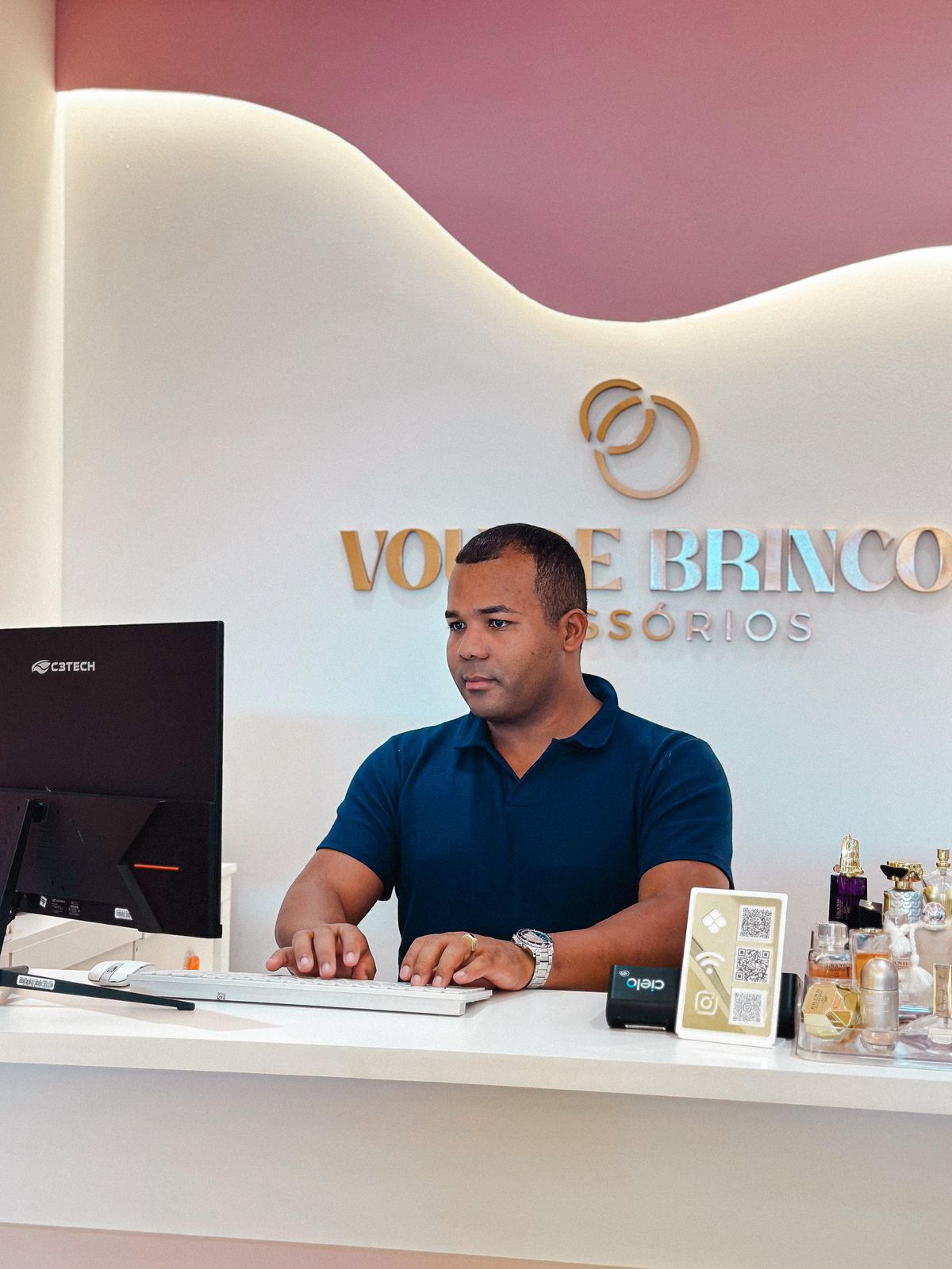 Mayconn trabalhando no computador atrás do balcão da loja Vou de Brinco Acessórios